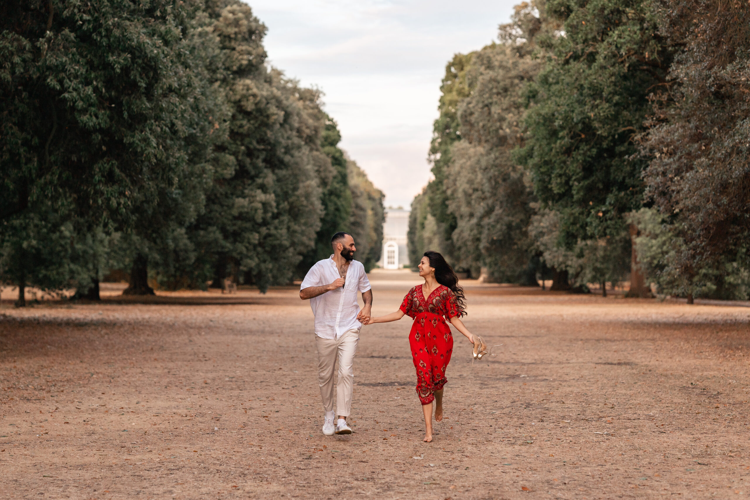 engaged couple, man wearing a white shirt, and woman wearing a red dress smile and run through the grounds of Kew Gardens in front of the Palm House