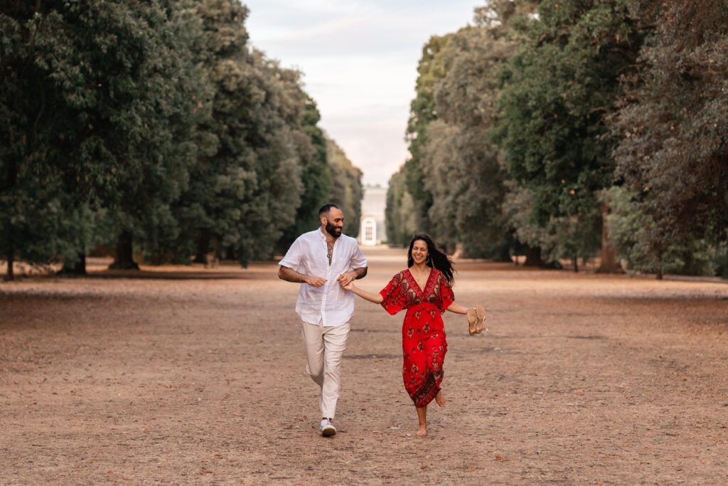 engaged couple, man wearing a white shirt, and woman wearing a red dress smile and run through the grounds of Kew Gardens in front of the Palm House