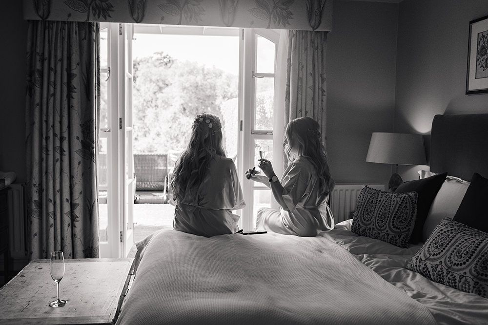 bridesmaids sit on edge of bed by window doing make up at Chewton Glen Wedding