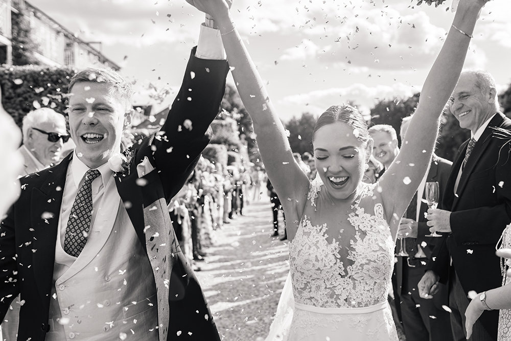 Bride and groom hold hands in the air in celebration whilst guests throw confetti