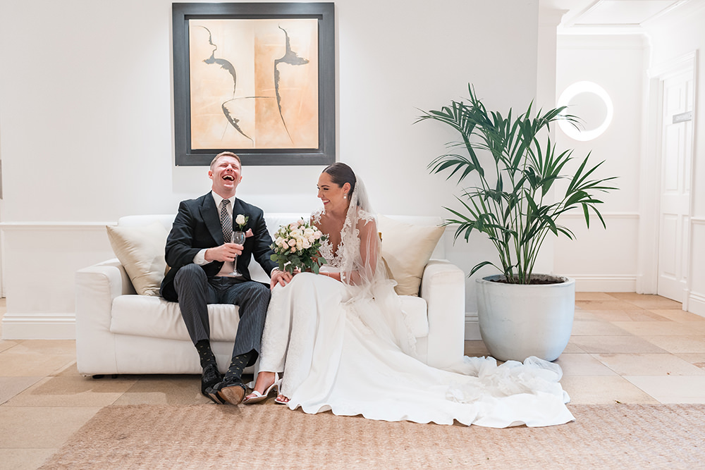 bride and groom sit on white sofa laughing at Chewton Glen Wedding