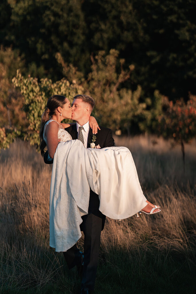 Groom carries bride through long grass in Chewton Glen apple orchard at sunset