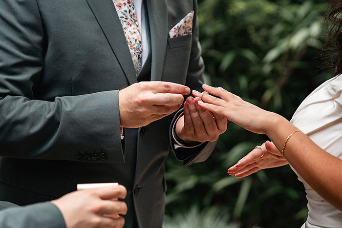 Groom placing ring on brides finger, behind them a forest of trees