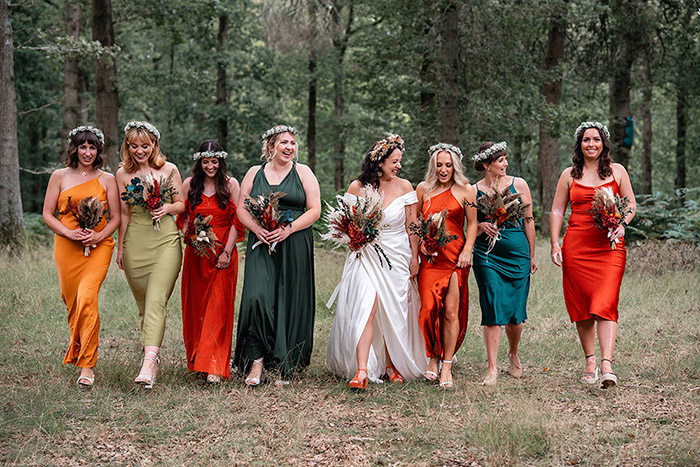 bride and bridesmaids stand together smiling in the forest, they are wearing jewel coloured dresses