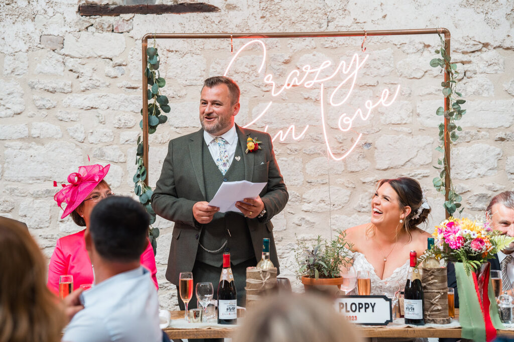 groom makes wedding speech infront of 'crazy in love' neon sign as bride laughs