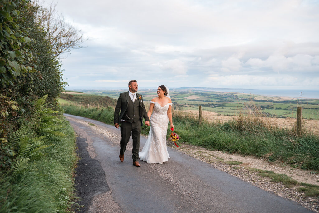 bride and groom walk hand in hand with Dorset hills and coastline behind them