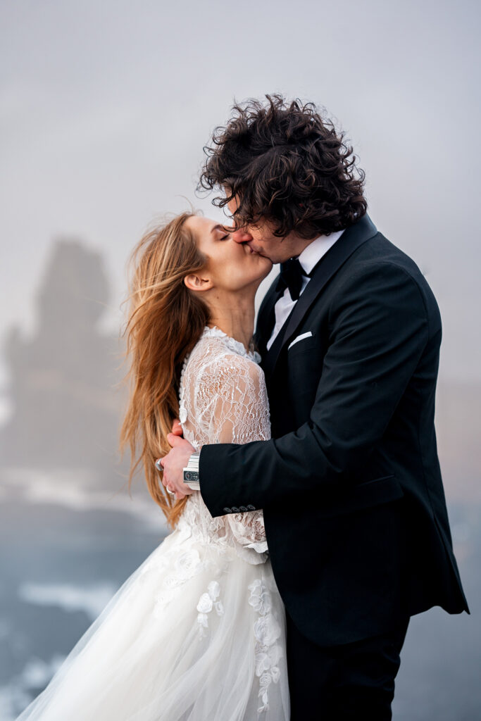bride and groom in tux and white wedding dress kiss on cliffs overlooking ocean in Iceland elopement