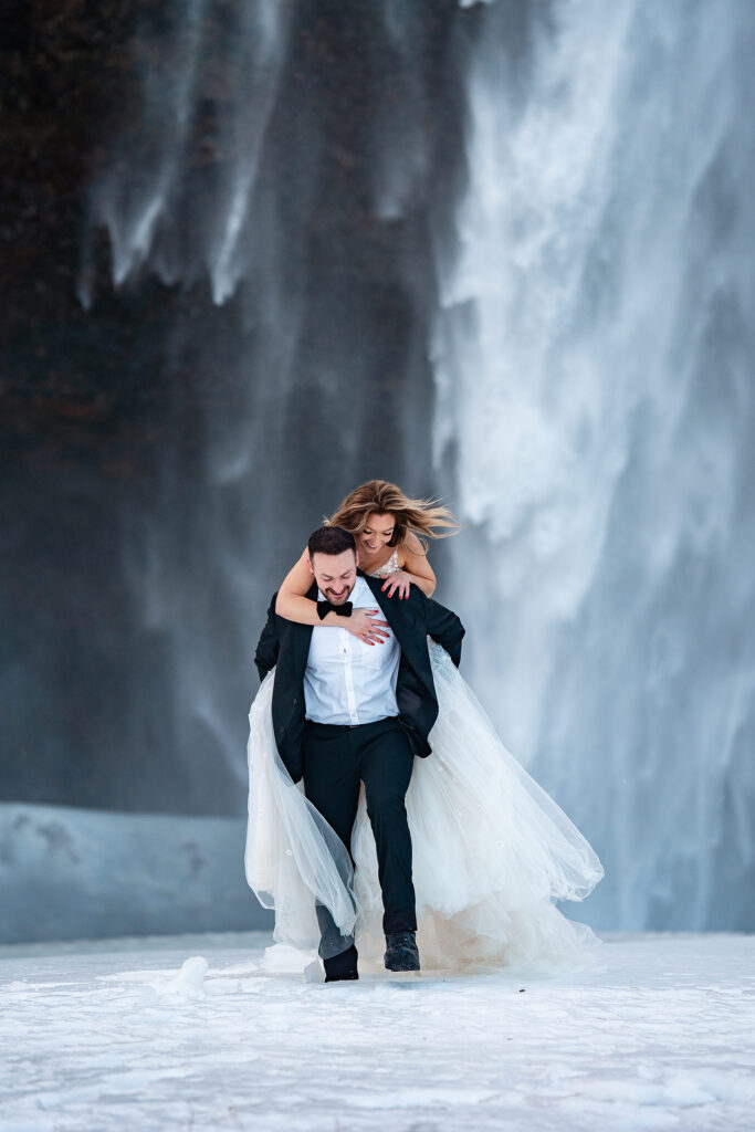 groom in Tux and Bride in Maramarie wedding dress, give piggy pack in front of Icelandic Waterfall
