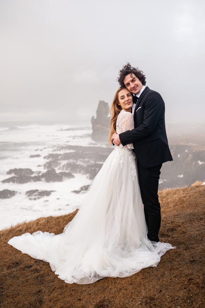bride and groom cuddle on rugged cliff overlooking ocean in Iceland