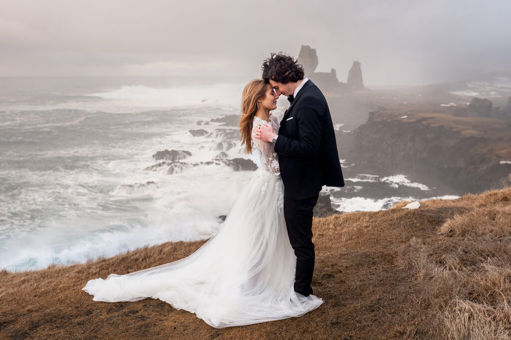 Newlywed couple embrace on a windy cliff edge in Iceland, overlooking the Ocean