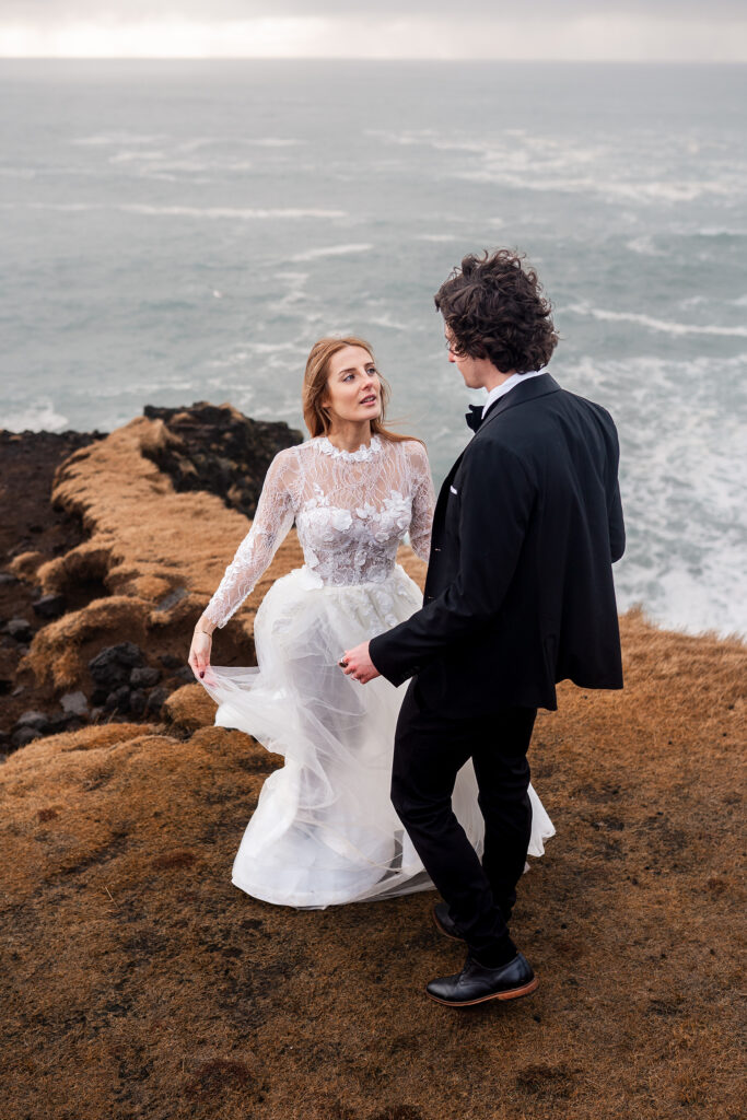 bride in white dress looks up at groom, both stood on cliff in Iceland