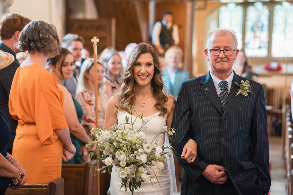 bride walking down the aisle in a church with her dad, smiling and holding white bouquet