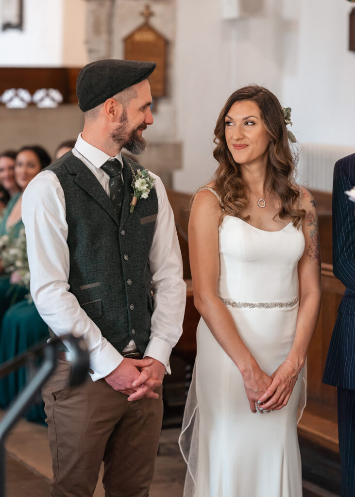 bride and groom give eachother a cheeky grin at the top of the aisle at a church wedding