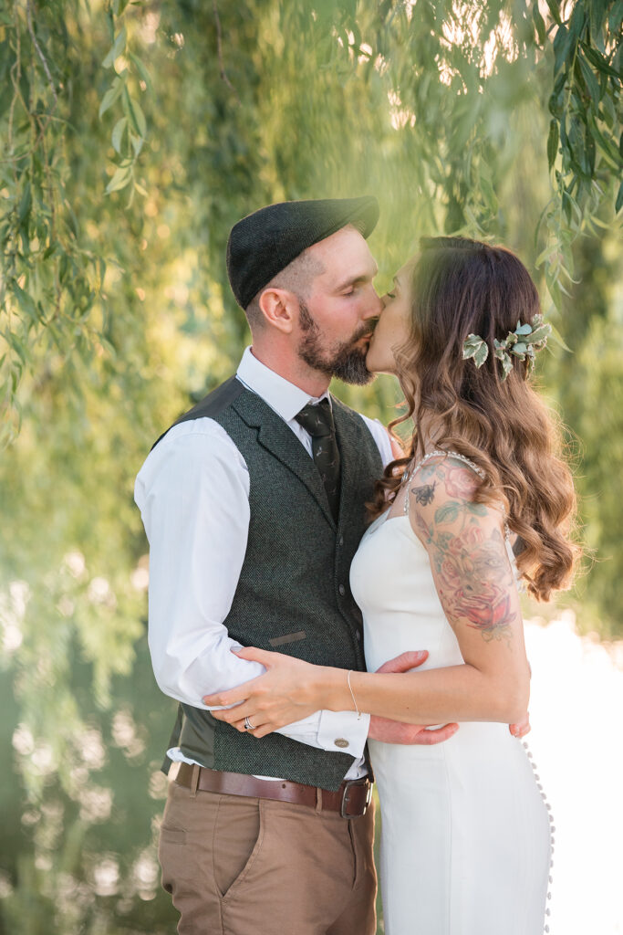 tattooed bride and groom wearing a cap kiss under a willow tree