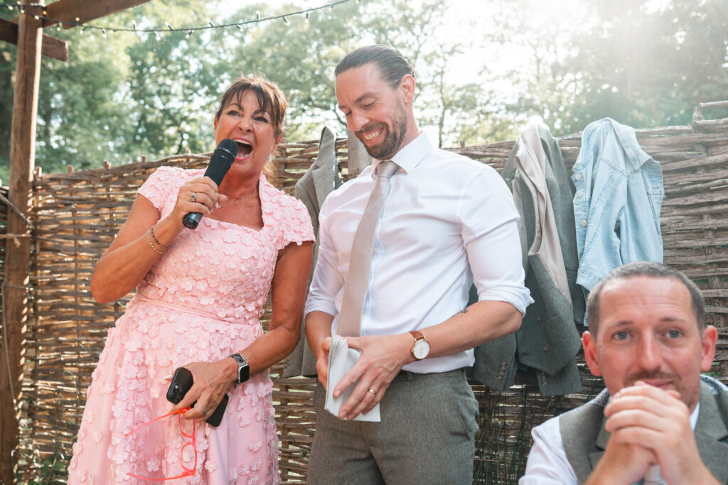Mother of the Bride stands next to groom holding microphone, both laughing