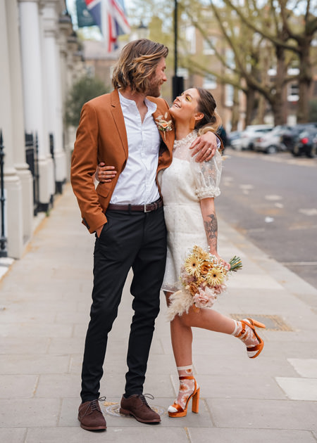 Bride wearing short dress and orange heels kisses groom in rust coloured suit