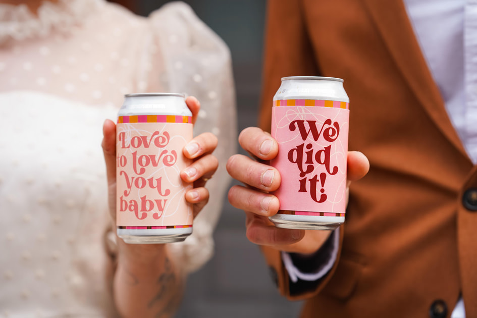 bride and groom hold personalised beer cans that say 'we did it'