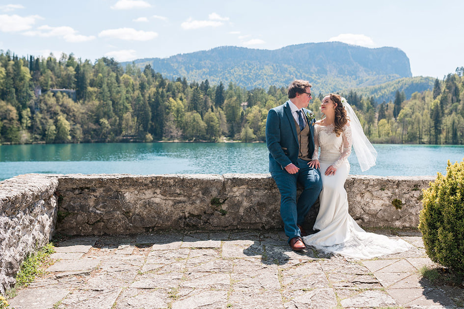 Bride and groom sit on wall overlooking Lake Bled