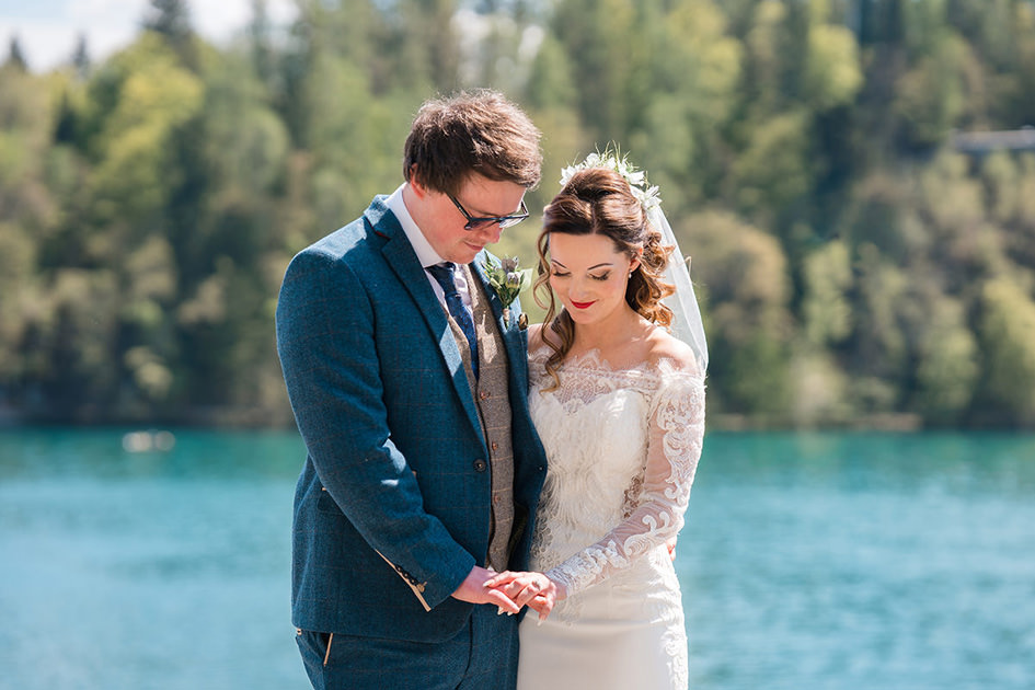 newly weds look down at wedding rings with Lake bled behind them